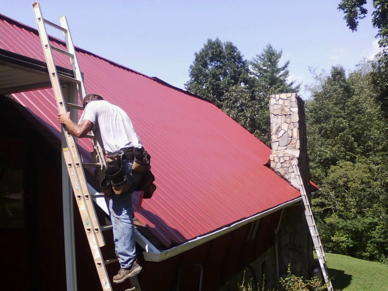 Roofer on a ladder beside a red metal roof and stone chimney — Soffit & Fascia Repair by Roofing Brothers in Ward